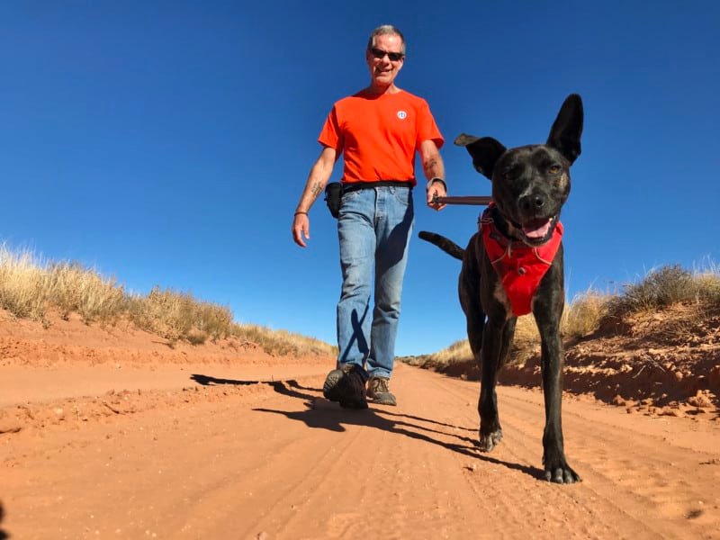 Man in orange t-shirt walking smiling dog on Willow Flats Road in Arches National Park - Moab, UT